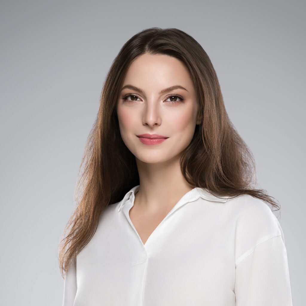 Studio headshot of a woman with long brown hair and a professional smile, wearing a white V-neck blouse. She is set against a neutral grey background, conveying a clean and approachable professional look.